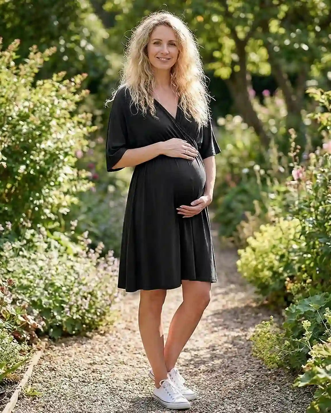 Pregnant woman in a black cross-front maternity midi dress with 3/4 sleeves, cradling her bump in a garden.