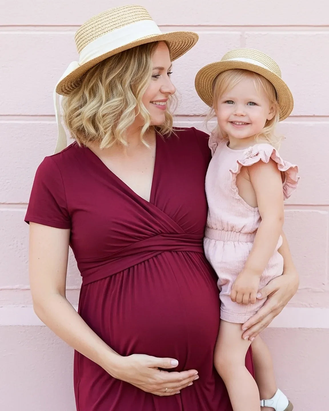 Pregnant woman in a burgundy Twist Front Draped Maternity Nursing Maxi Dress holding a child, both wearing straw hats.