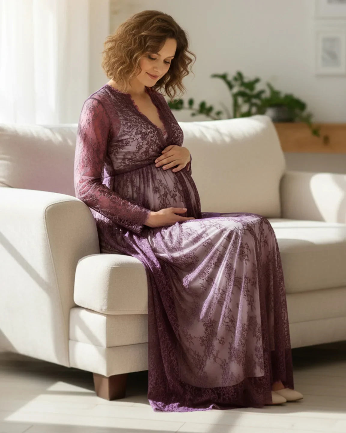 Pregnant woman in a sheer floral lace empire waist maternity maxi dress, deep purple, sitting on a white sofa.