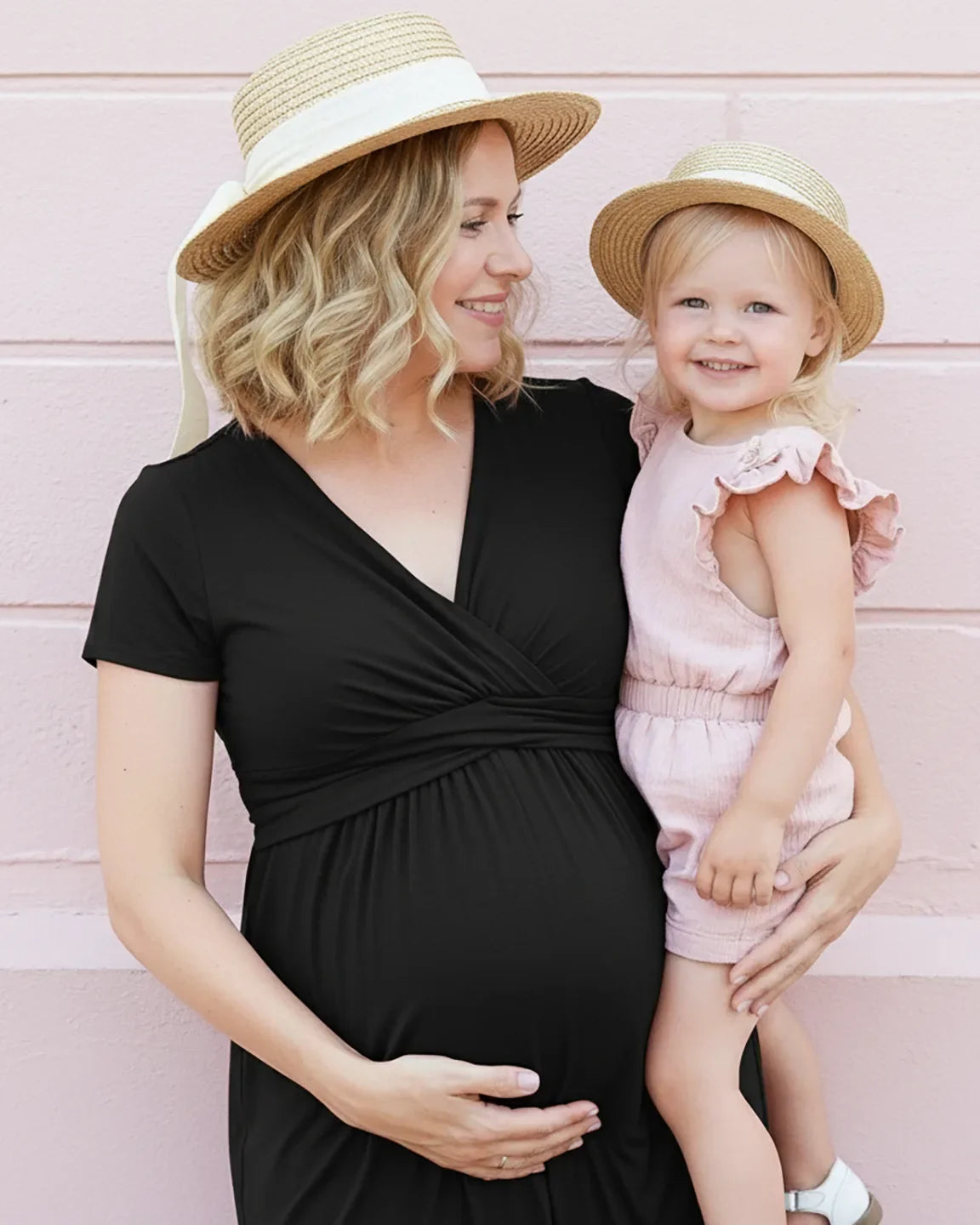 Pregnant woman in a black twist-front draped nursing maxi dress, holding a child, both wearing straw hats.