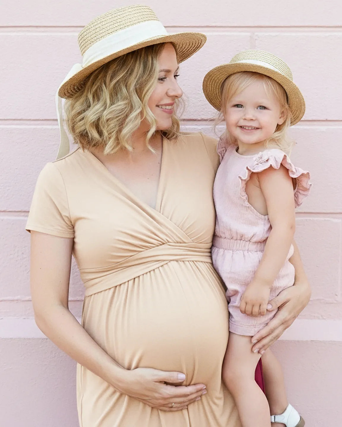 Pregnant woman in a beige twist-front draped nursing maxi dress, holding a toddler in a pink romper.