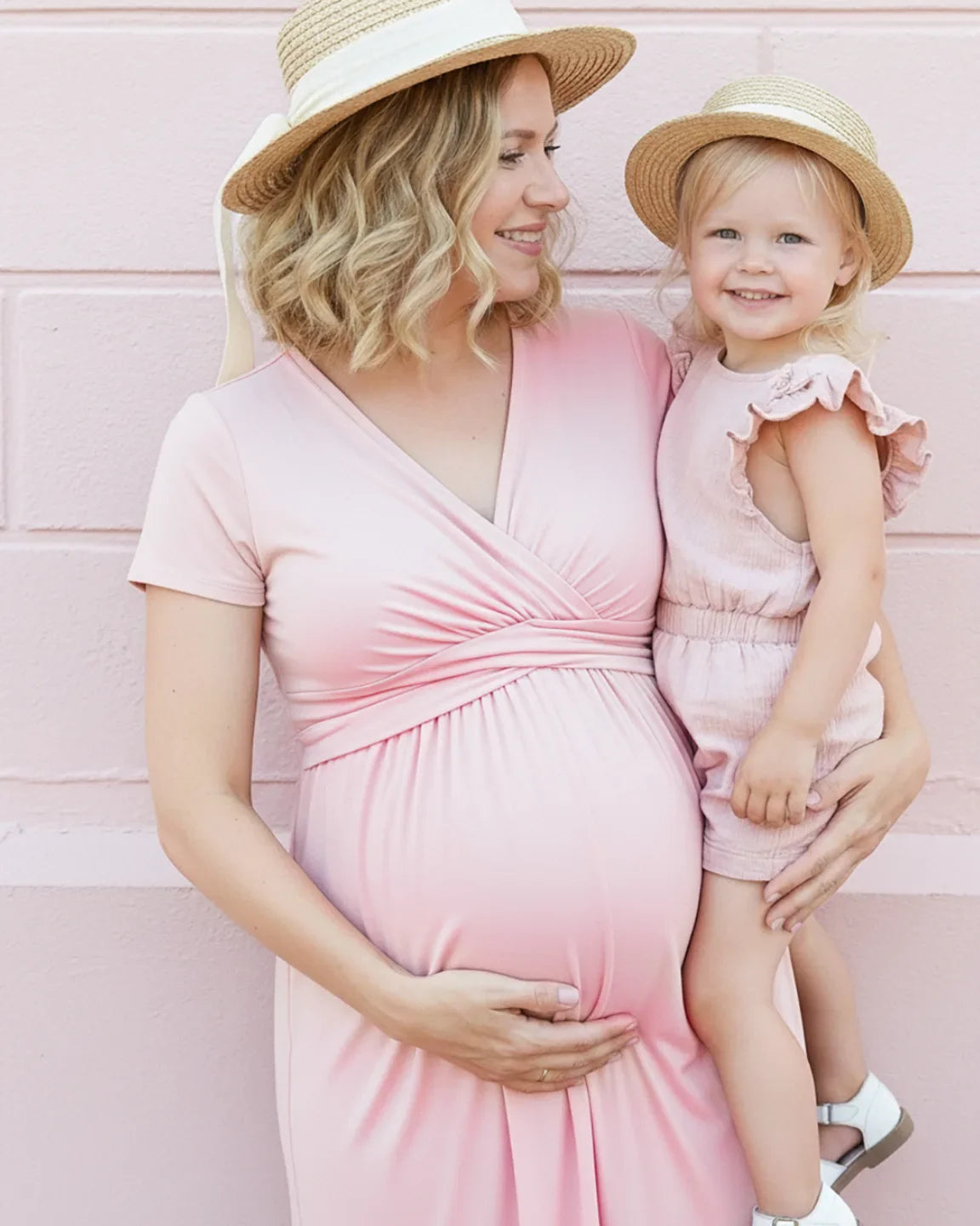 Pregnant woman in a pink Twist Front Draped Maternity Nursing Maxi Dress holding a child, both wearing straw hats.