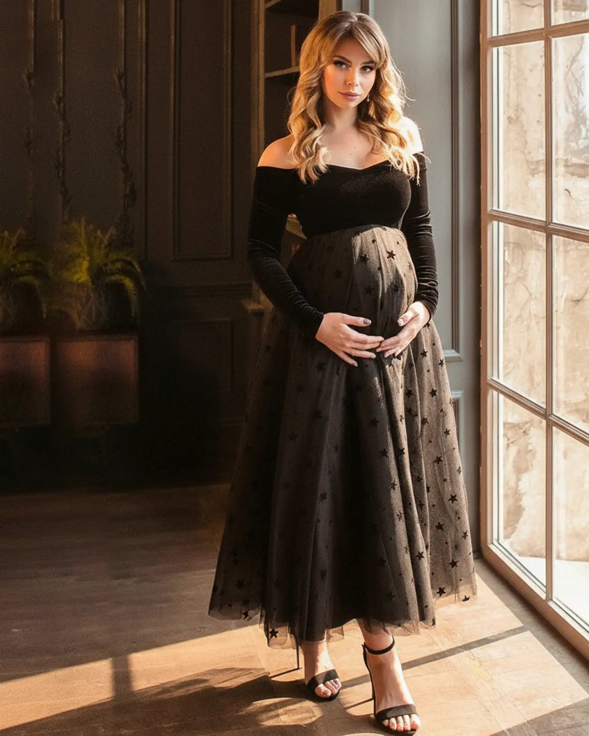 Maternity midi dress: black off-shoulder velvet bodice, star-flocked mesh skirt, woman cradling bump.