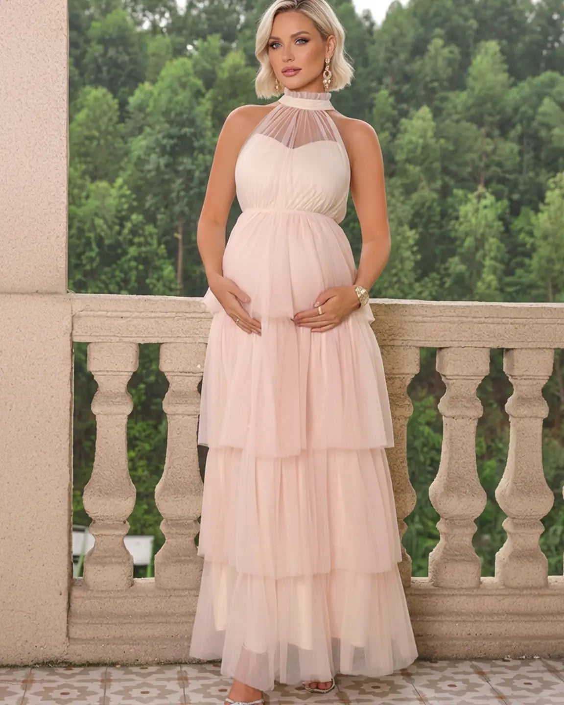 Blonde pregnant woman in a blush pink halter neck tiered mesh maternity gown on a balcony.