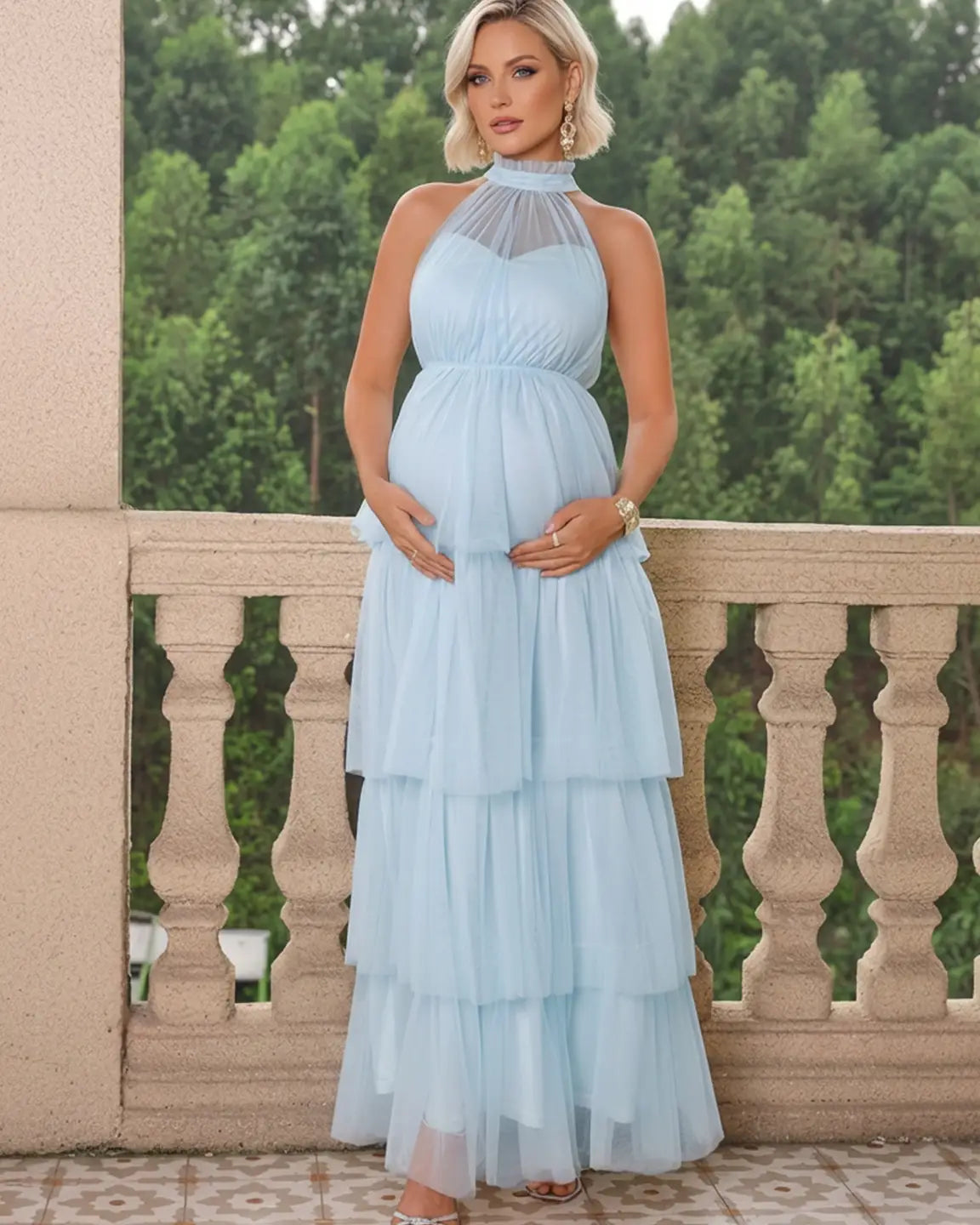 Maternity model in a light blue halter neck tiered mesh gown, holding her bump on a balcony.