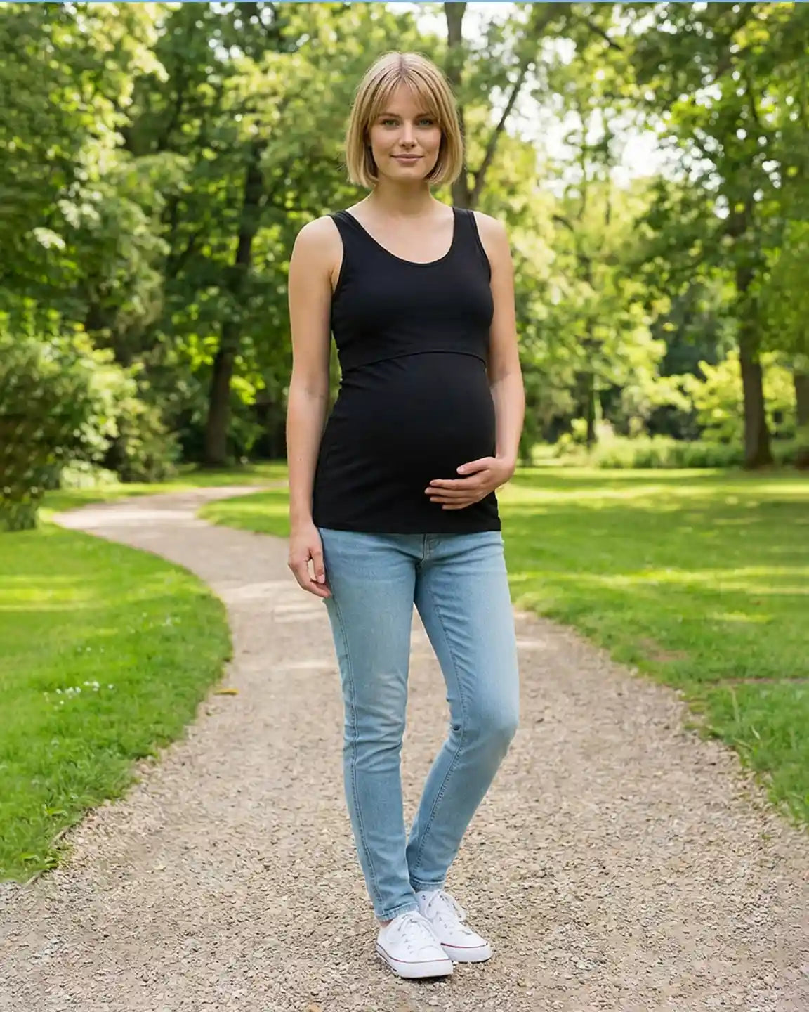 Pregnant woman in black stretch nursing-access maternity tank top and jeans, standing on a park path.
