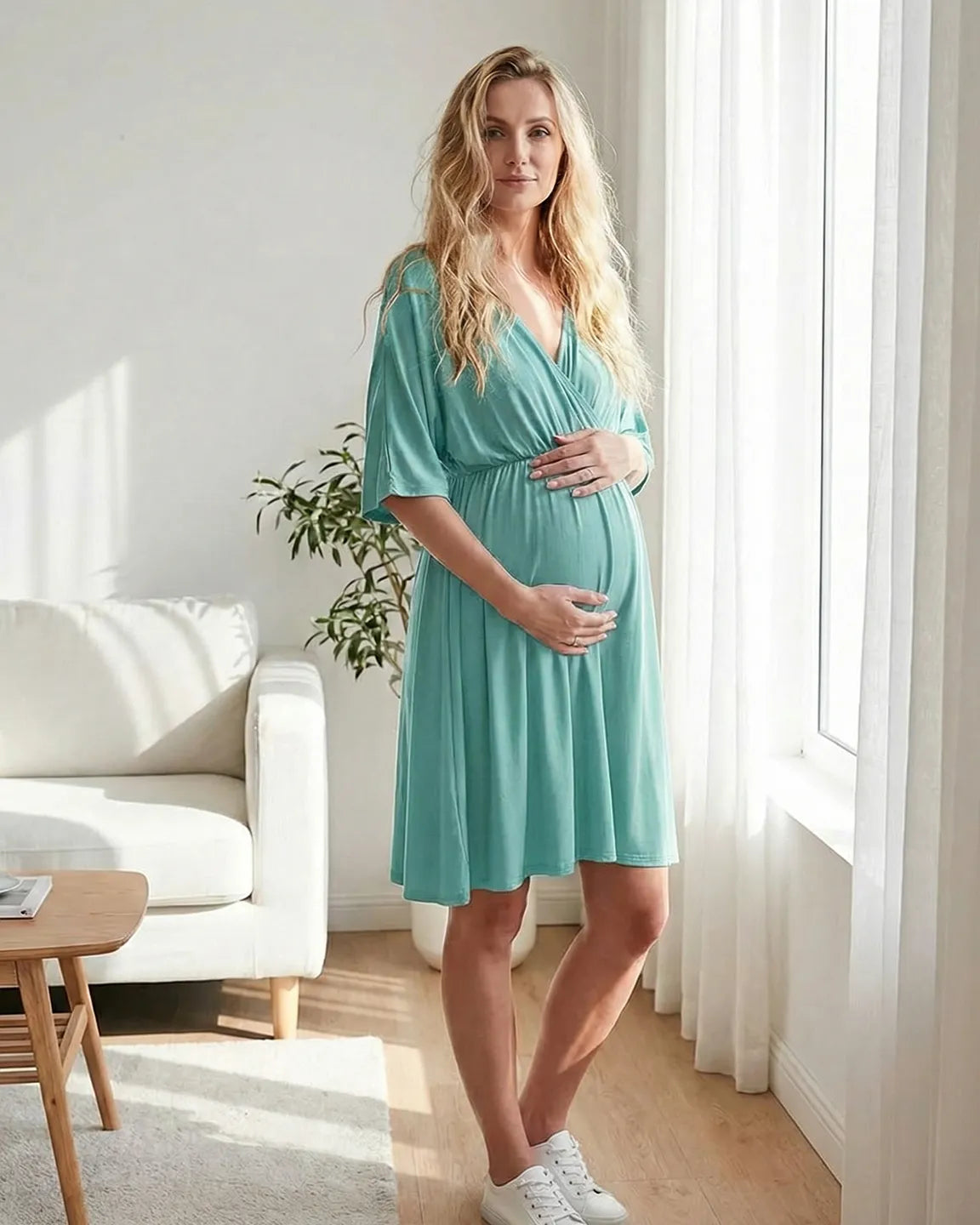 Blonde pregnant woman in a teal cross-front maternity midi dress with 3/4 sleeves, holding her bump.