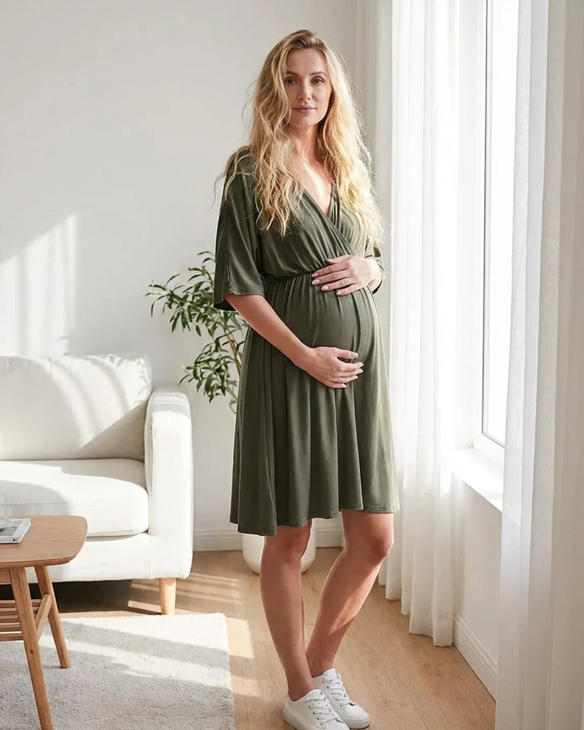 Blonde pregnant woman in a dark olive green cross-front maternity midi dress with 3/4 sleeves, holding her bump.