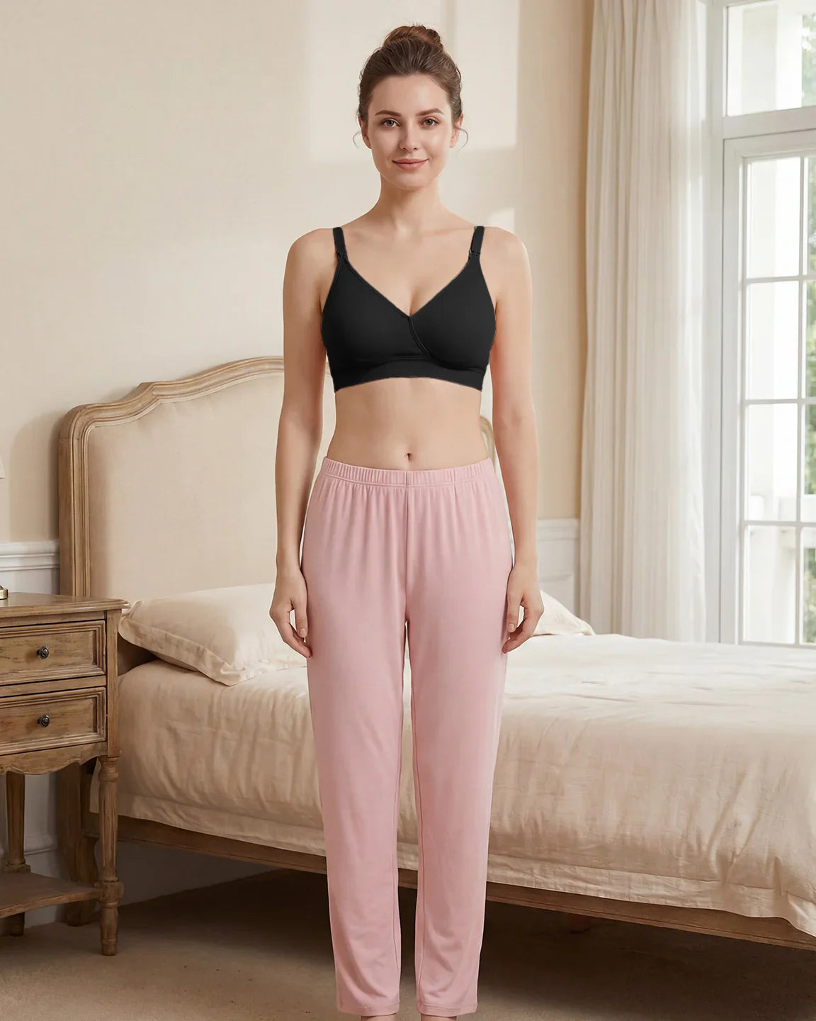 Woman in a black seamless cross-front wireless nursing bra and pink lounge pants, standing in a bedroom.