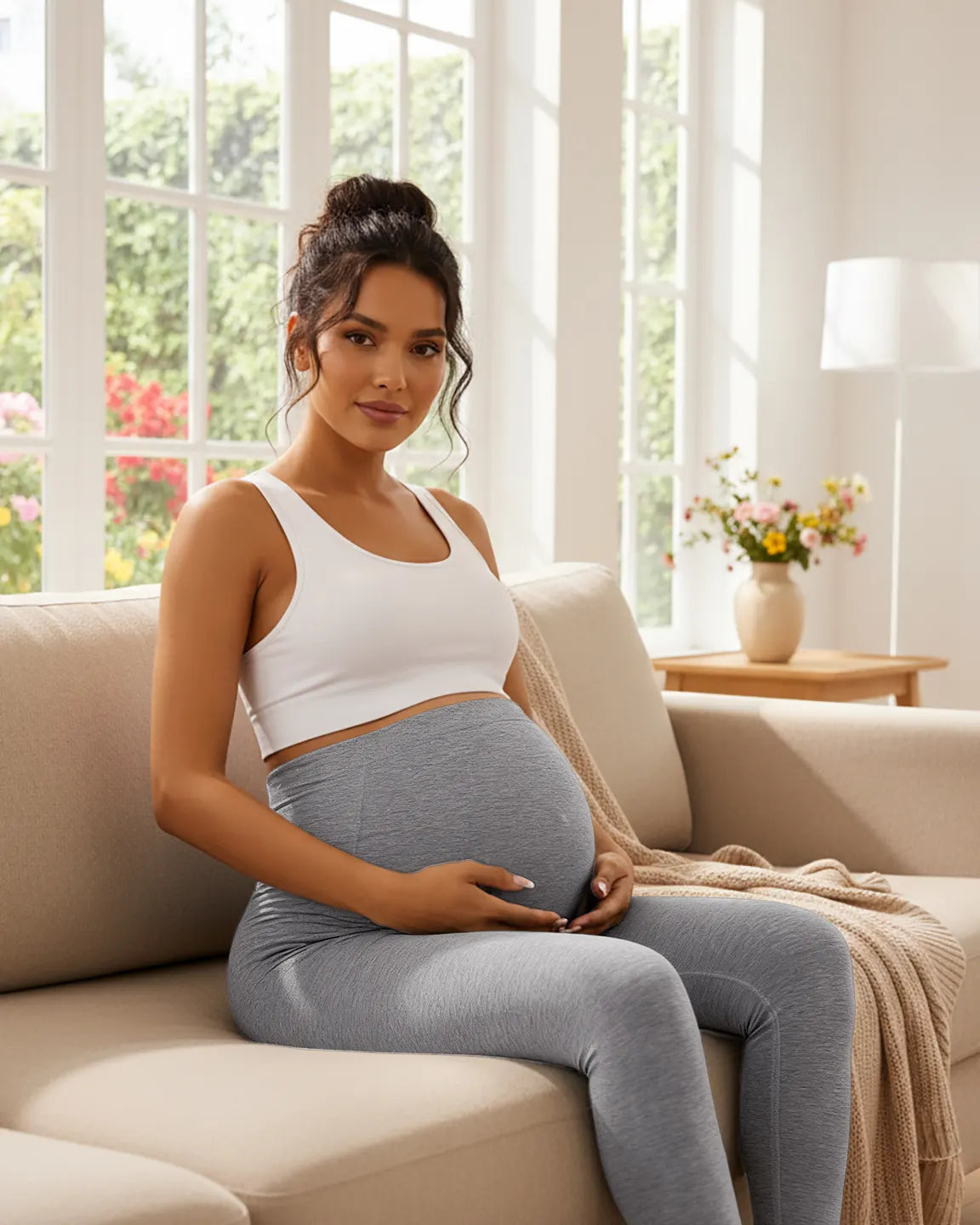 Pregnant woman in light gray seamless M-shape maternity leggings and white crop top, cradling her bump on a sofa.