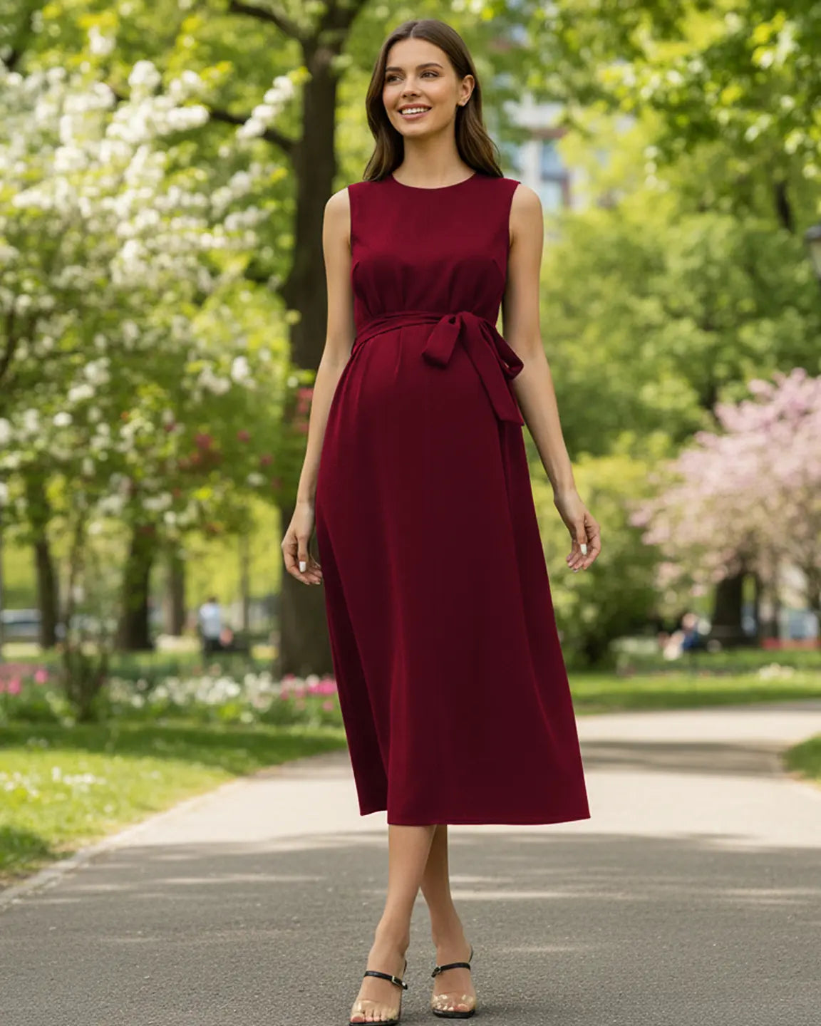 Smiling pregnant woman in a sleeveless burgundy midi dress with a tie-waist, walking outdoors.