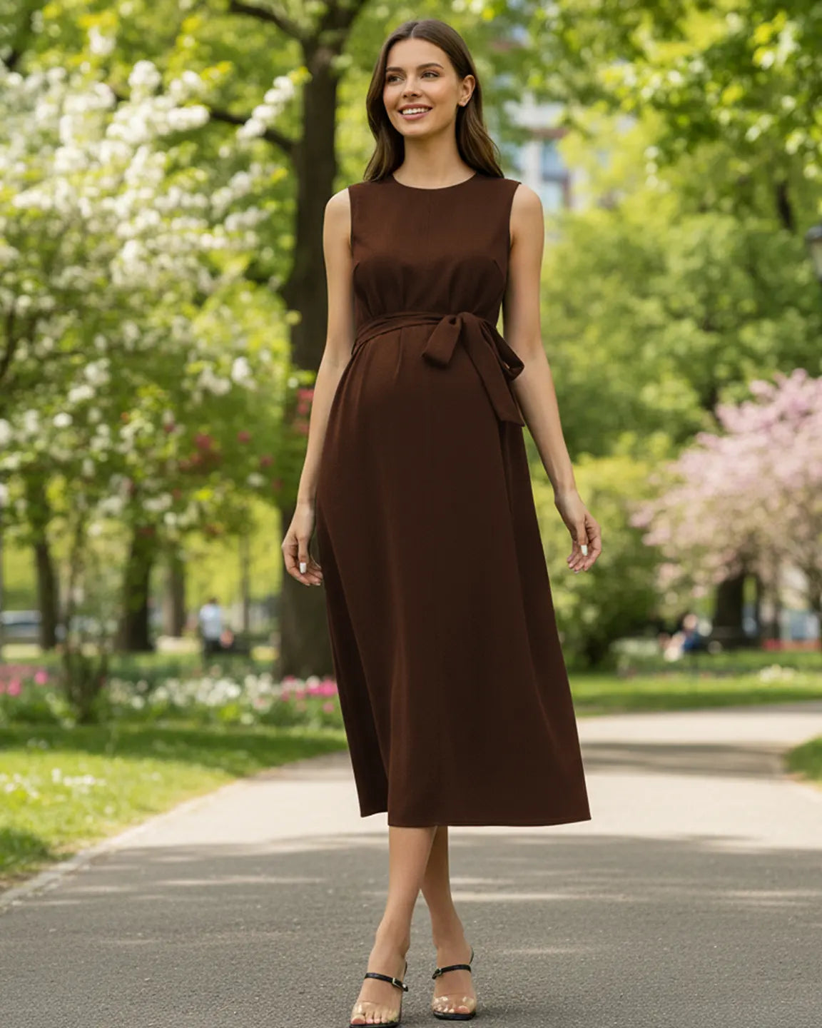Smiling pregnant woman in a brown sleeveless midi dress with a tie-waist, walking outdoors.