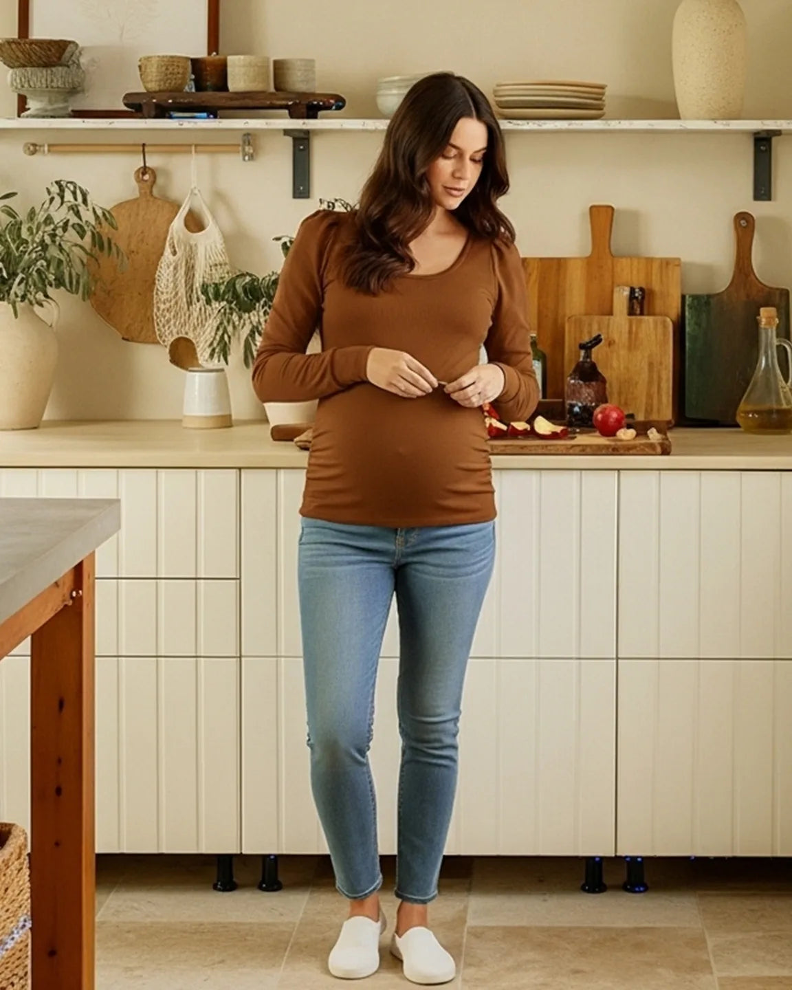Pregnant woman in a warm brown, long-sleeve ruched maternity top, light wash jeans, and white shoes.