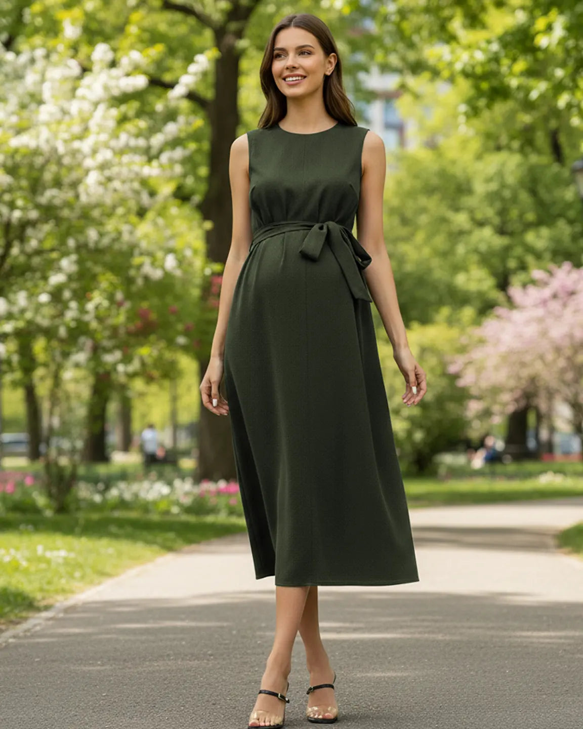 Smiling pregnant woman in a dark green sleeveless maternity midi dress with a tie-waist, walking in a park.