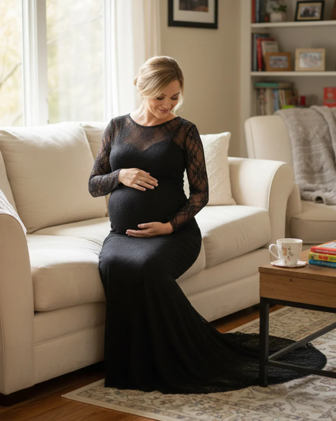 Pregnant woman in a black sheer lace trumpet maternity gown with long sleeves, sitting on a cream sofa.