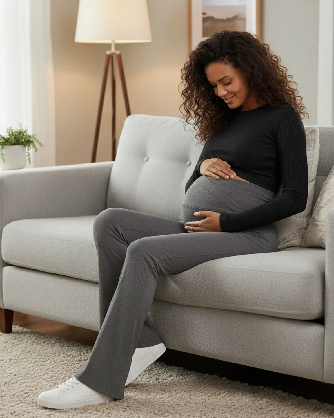 Smiling pregnant woman in charcoal gray crossed waistband flared maternity pants and a black top, sitting on a couch.