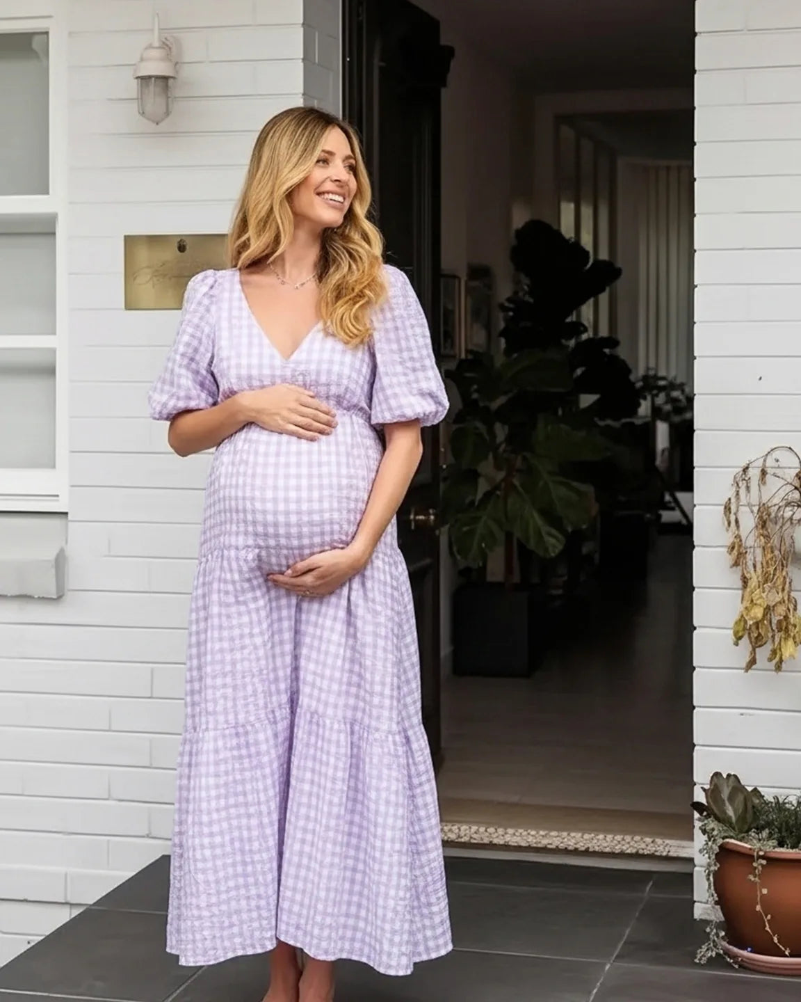 Smiling pregnant woman in a lavender gingham puffed sleeve maternity maxi dress, hands on bump.