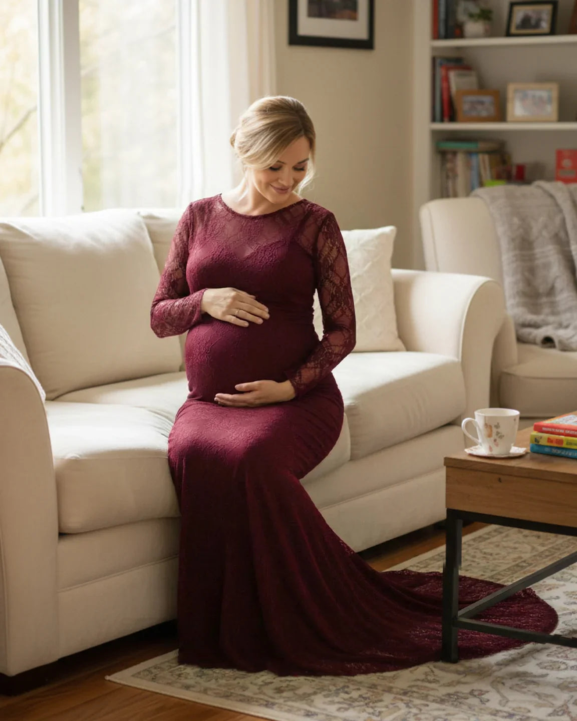 Pregnant woman in a long-sleeved, burgundy sheer lace trumpet maternity gown, sitting on a white sofa.