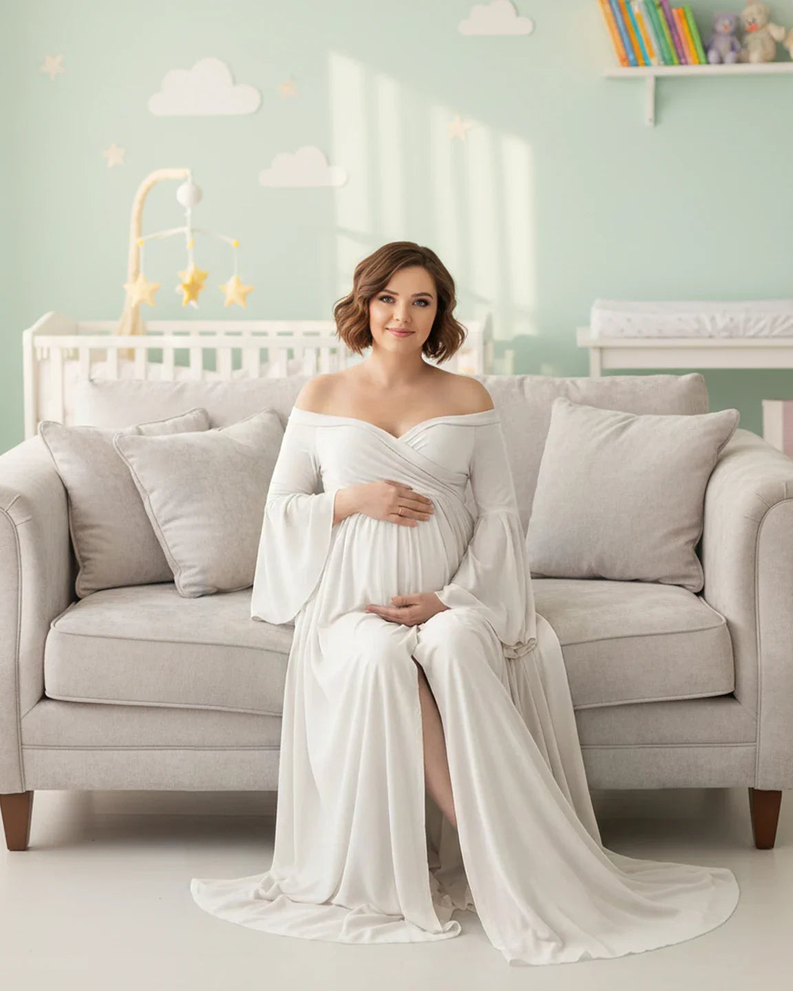 Pregnant woman in a white off-shoulder bell-sleeve maternity gown with a long train, sitting on a sofa.