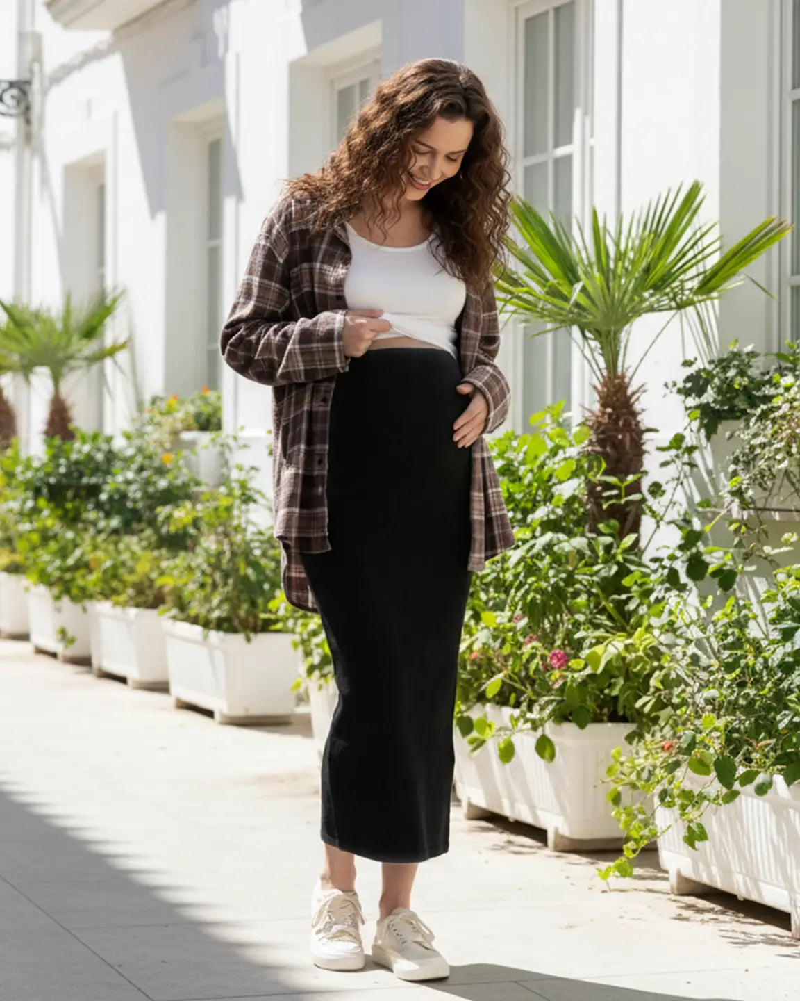 Smiling pregnant woman in a black midi skirt, white crop top, and brown plaid shirt, touching her bump.