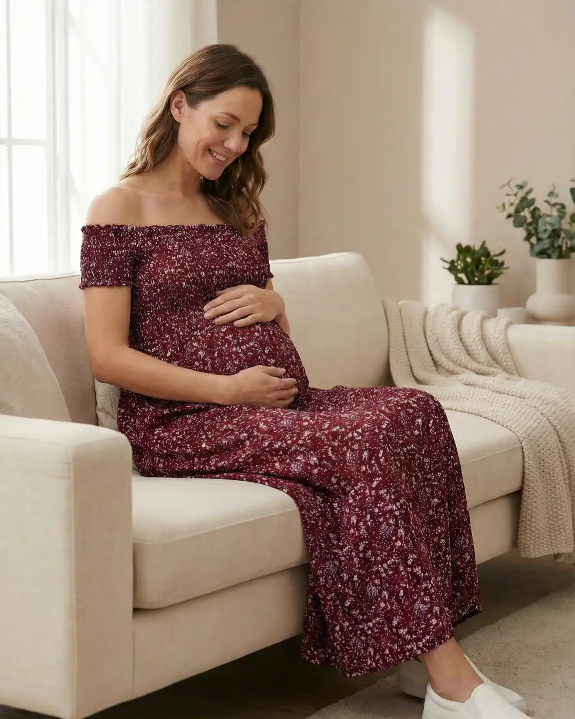 Smiling pregnant woman in a red floral off-shoulder maternity maxi dress with side slit, sitting on a sofa.