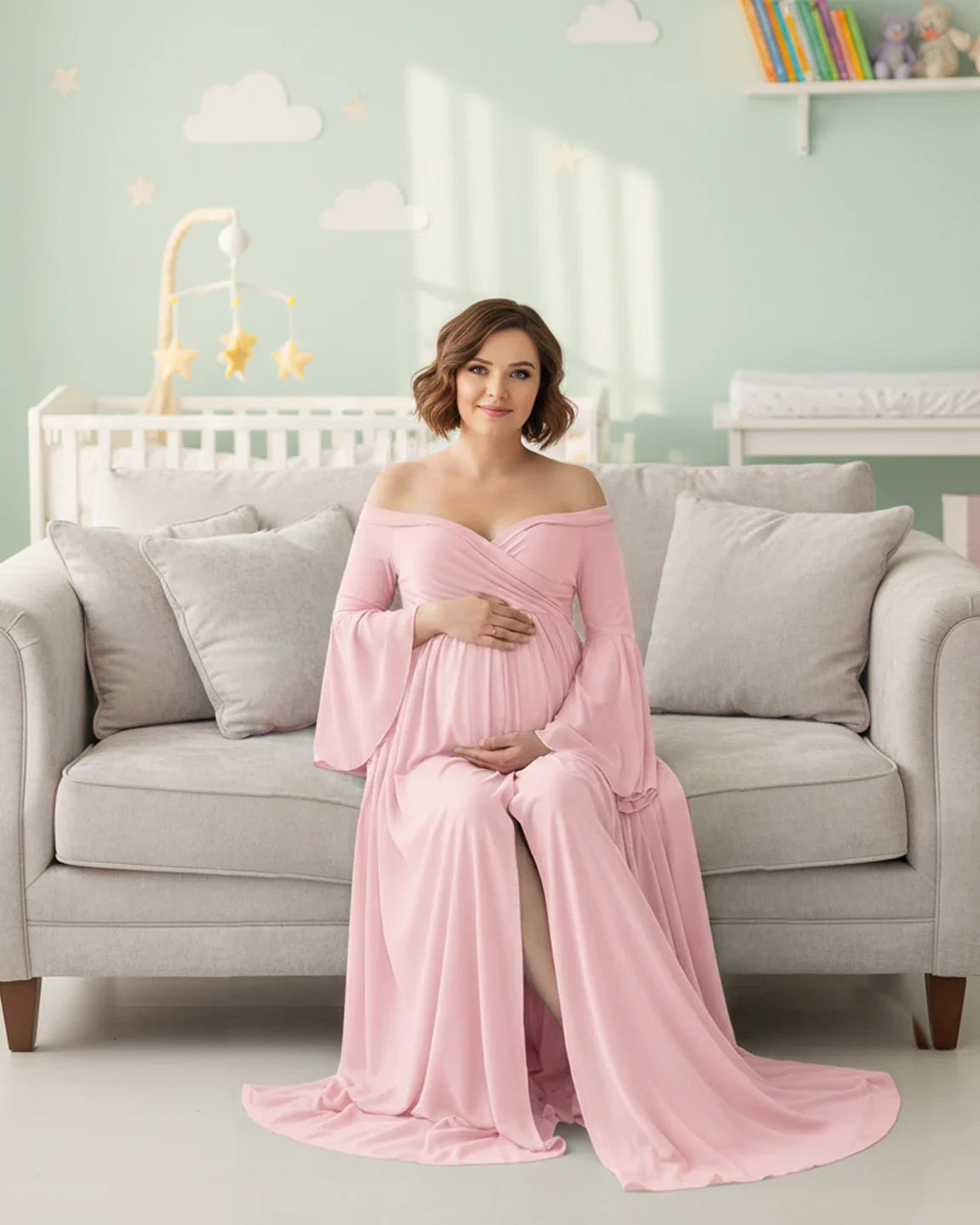 Pregnant woman in a pink off-shoulder bell-sleeve maternity gown with a long train, sitting on a sofa.