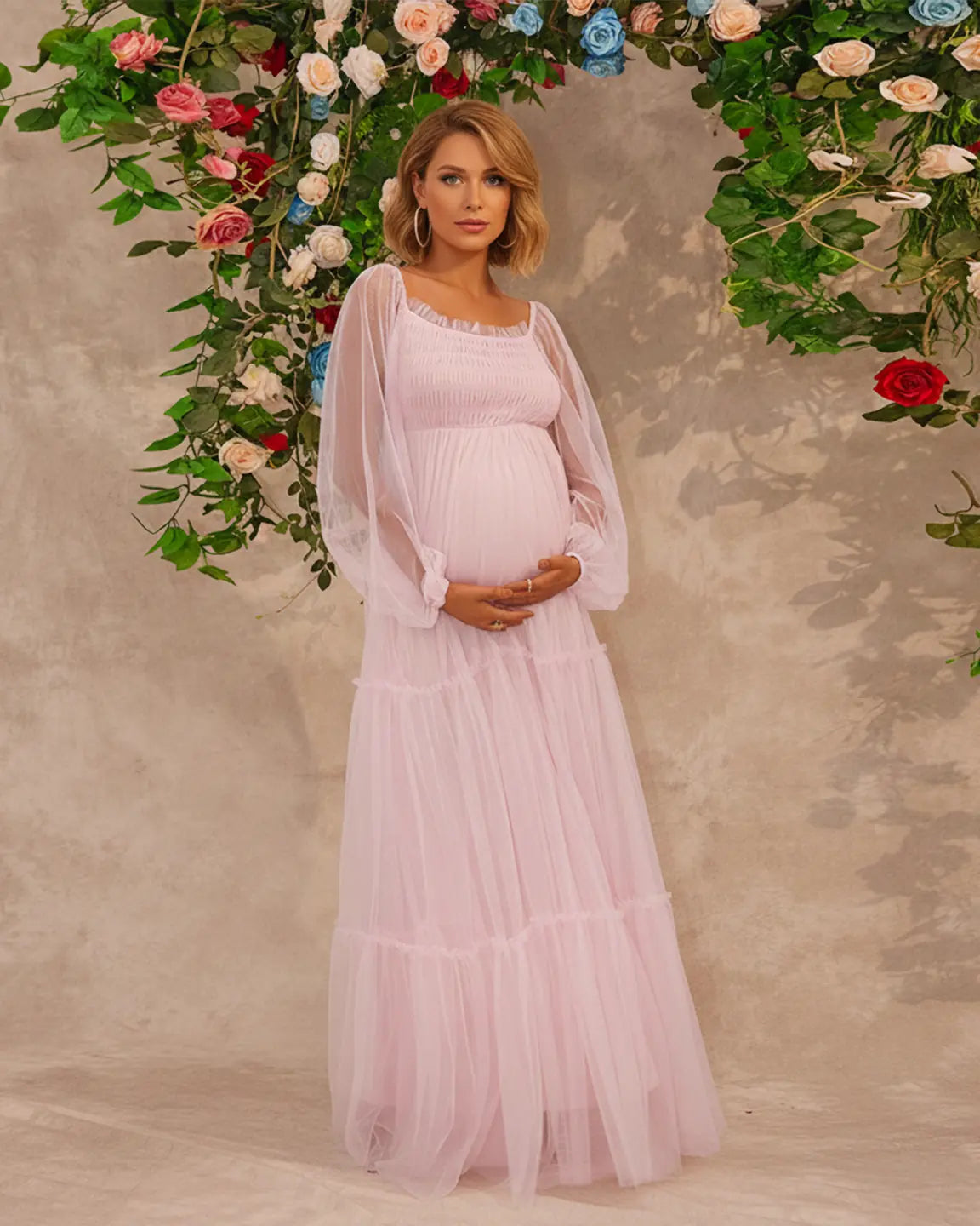 Pregnant woman in a pink tiered tulle maxi gown with lantern sleeves, standing under a floral arch.