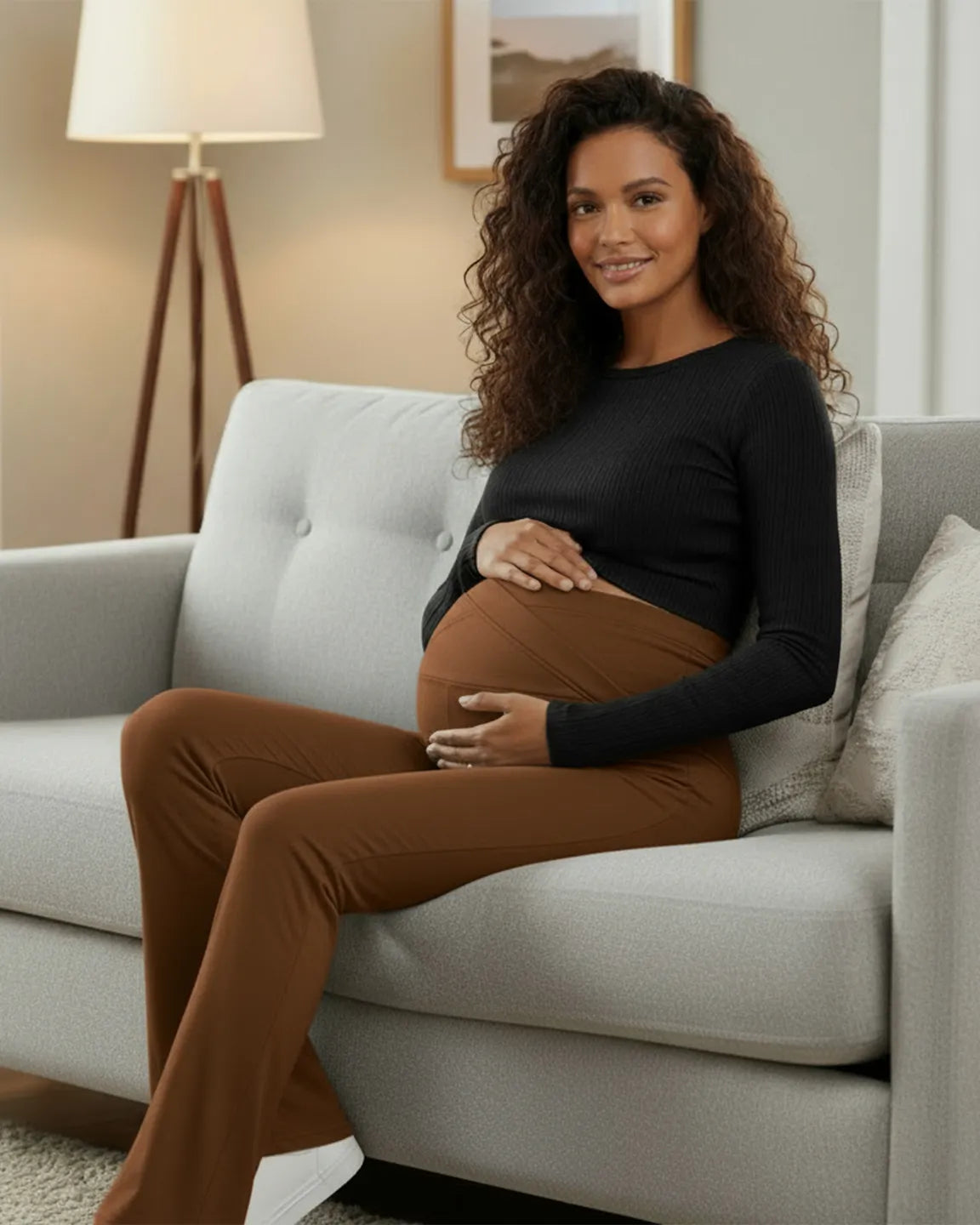 Smiling pregnant woman wearing brown crossed waistband flared maternity pants and a black top, sitting on a gray couch.