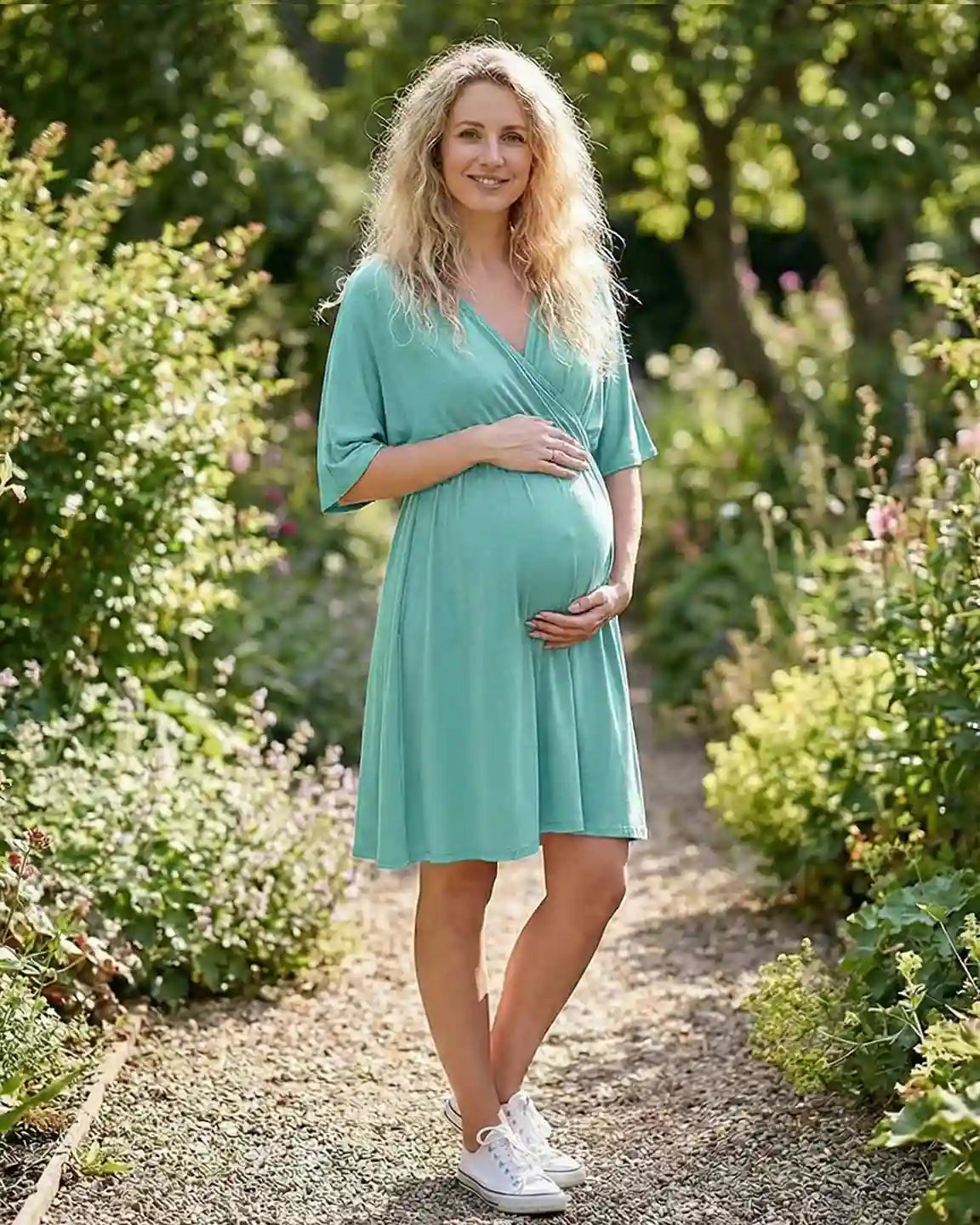 Pregnant woman in a teal cross-front midi dress with 3/4 sleeves, holding her bump in a garden.