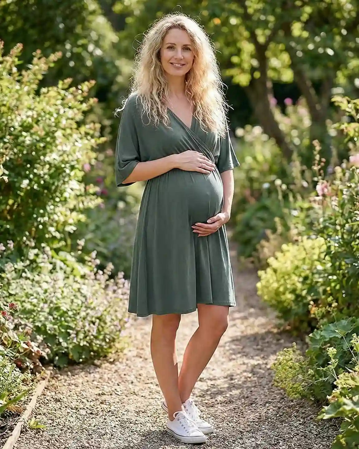 Pregnant woman in a soft olive green cross-front maternity midi dress with elbow-length sleeves, holding her bump in a garden.