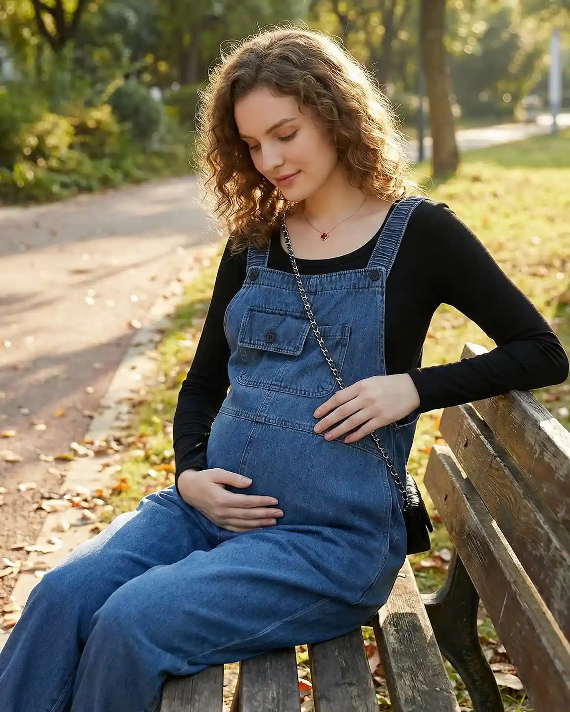 Pregnant woman in loose-fit blue denim maternity overalls with adjustable straps and pockets, sitting on a park bench.