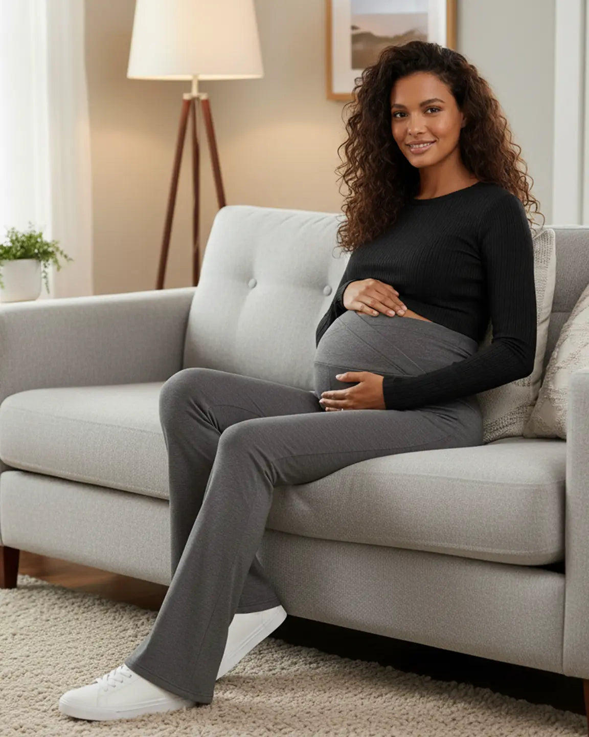 Smiling pregnant woman in charcoal gray crossed waistband flared maternity pants and a black top, sitting on a sofa.