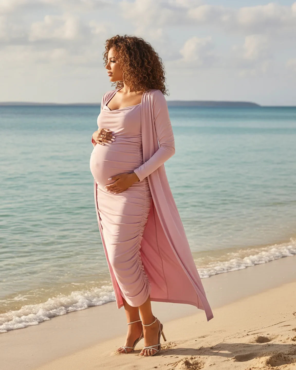 Pregnant woman in a pink side-ruched slip dress and long cardigan on a beach.