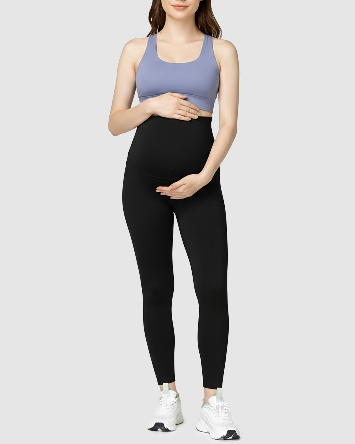 Woman in black high-waist soft stretch maternity leggings and a purple sports bra, cradling her bump.