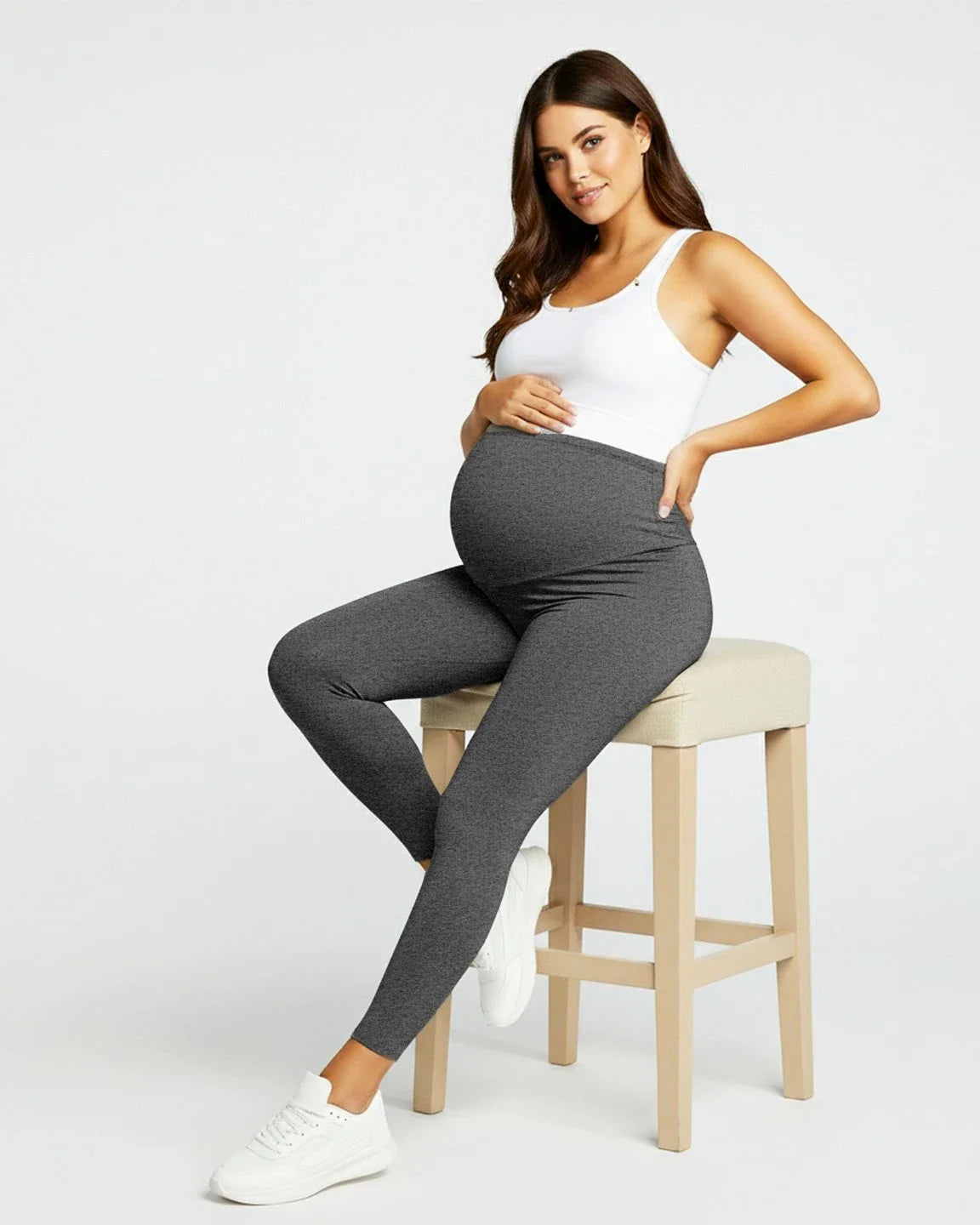 Pregnant woman in dark gray sculpting ankle-length maternity leggings and a white tank top, sitting on a stool.