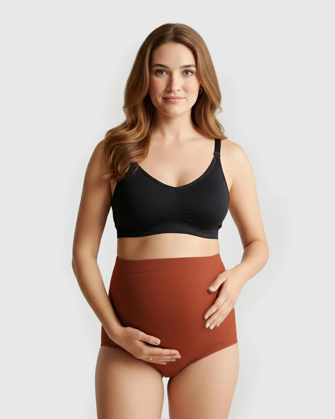 Pregnant woman in a black bra and rust-colored high-waist seamless maternity panties, cradling her bump.