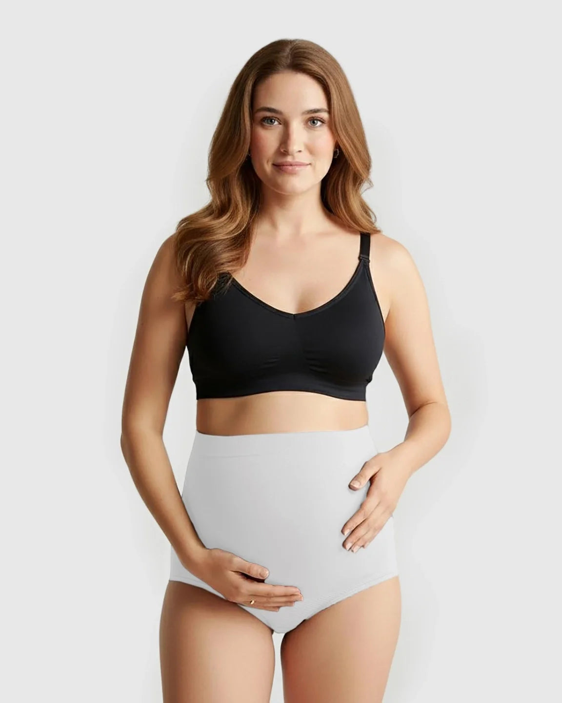 Pregnant woman wearing light grey seamless high-waist maternity panties and a black bra, holding her bump.