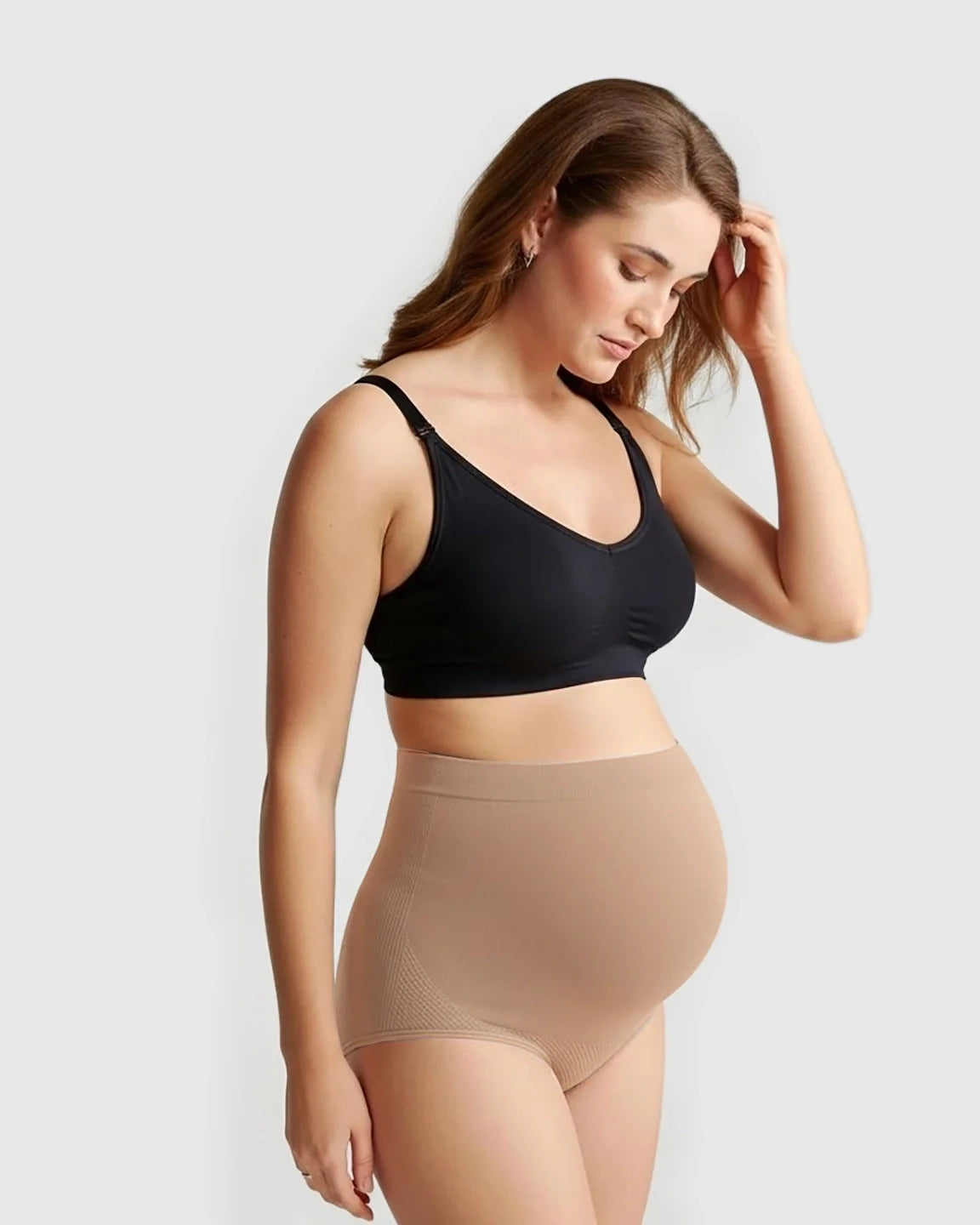 Pregnant woman modeling Bump Society high-waist seamless maternity panties in nude, with a black bra.