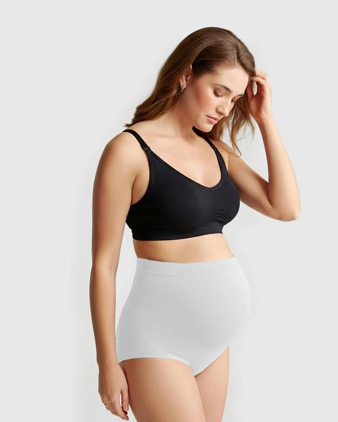 Pregnant woman wearing white high-waist seamless maternity panties and a black bra, looking down.