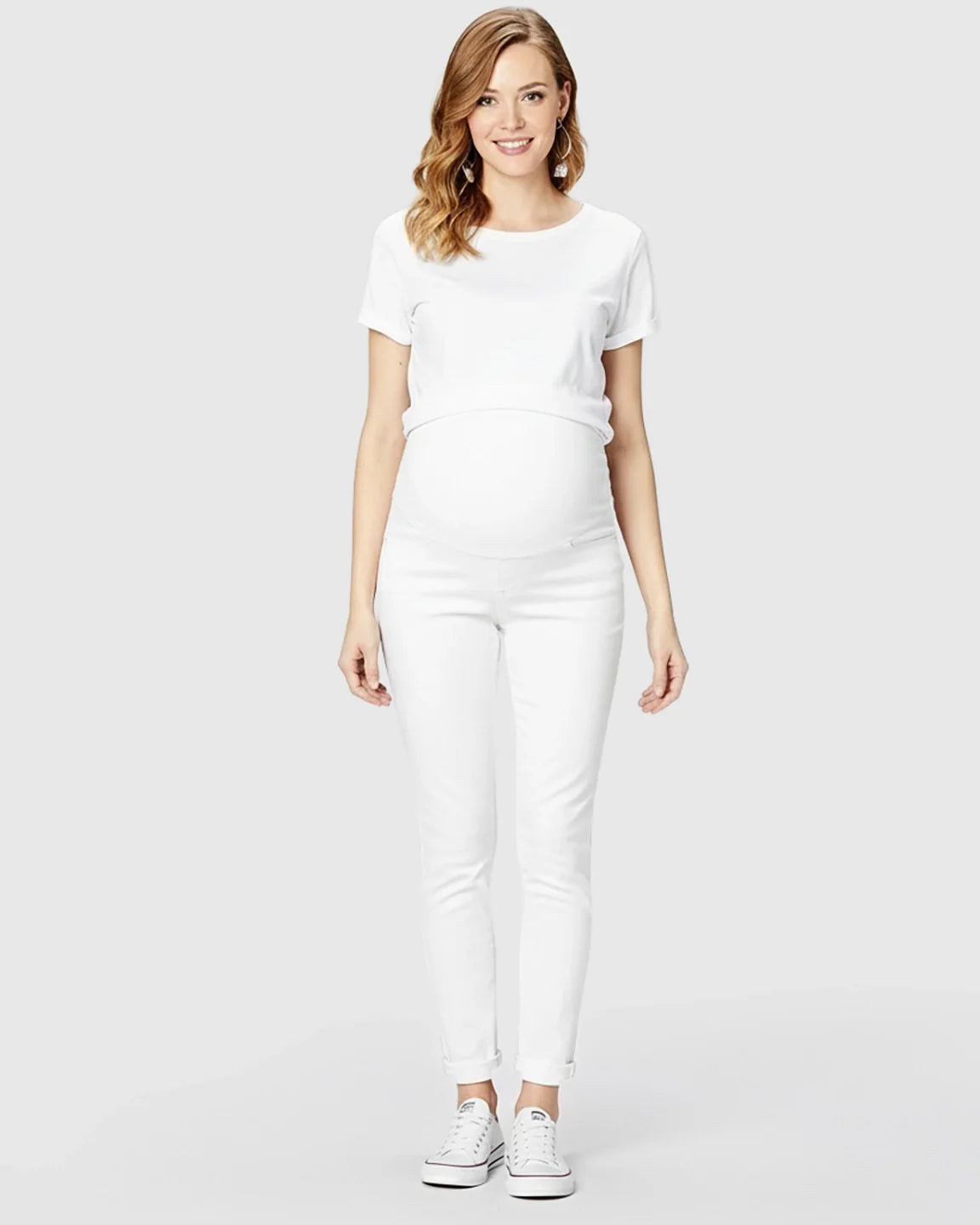 Smiling pregnant woman wearing a white t-shirt and white full panel maternity skinny jeans with cuffed hems.