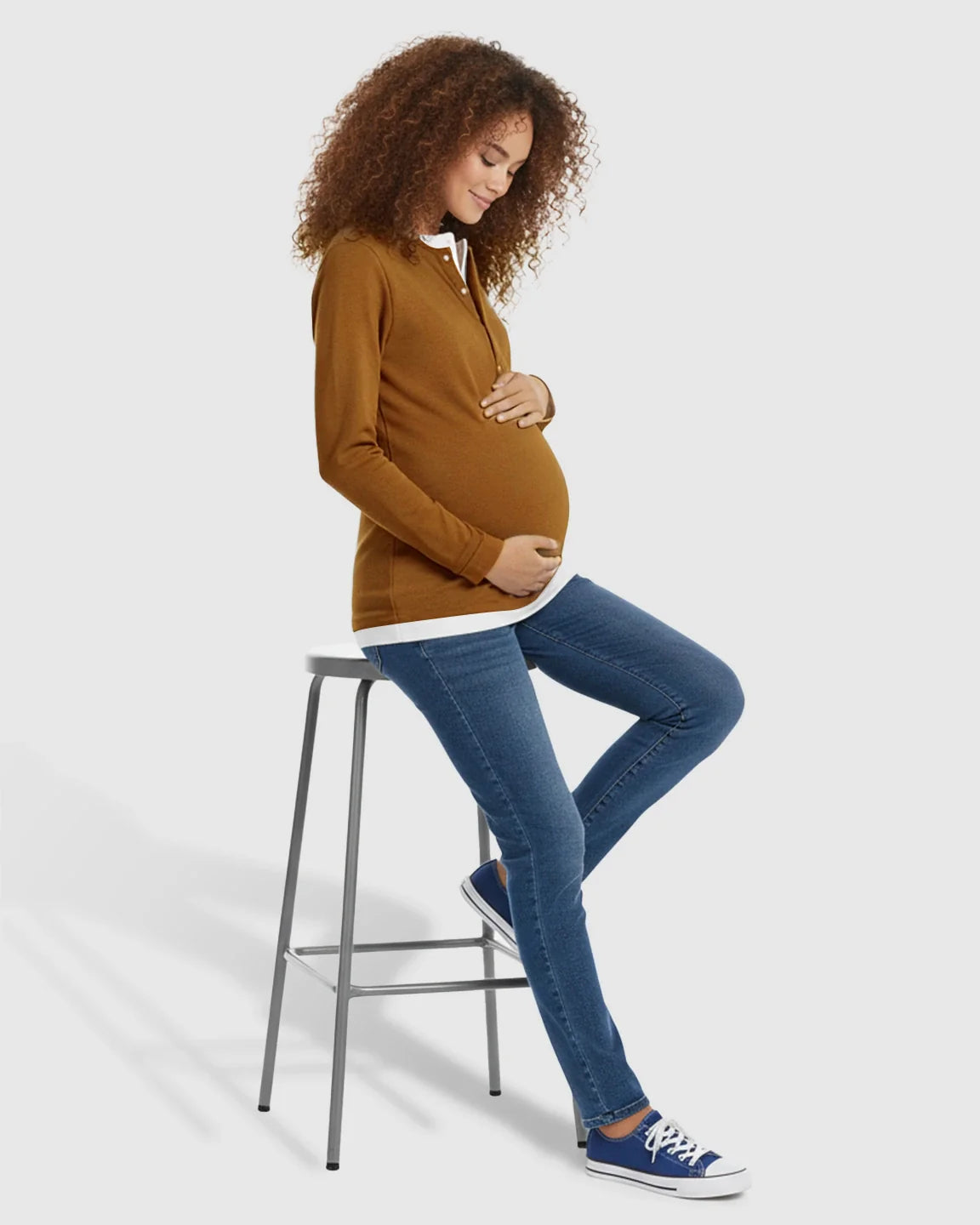 Smiling pregnant woman in a brown button-front henley maternity and nursing top with a white underlayer, blue jeans, and sneakers.