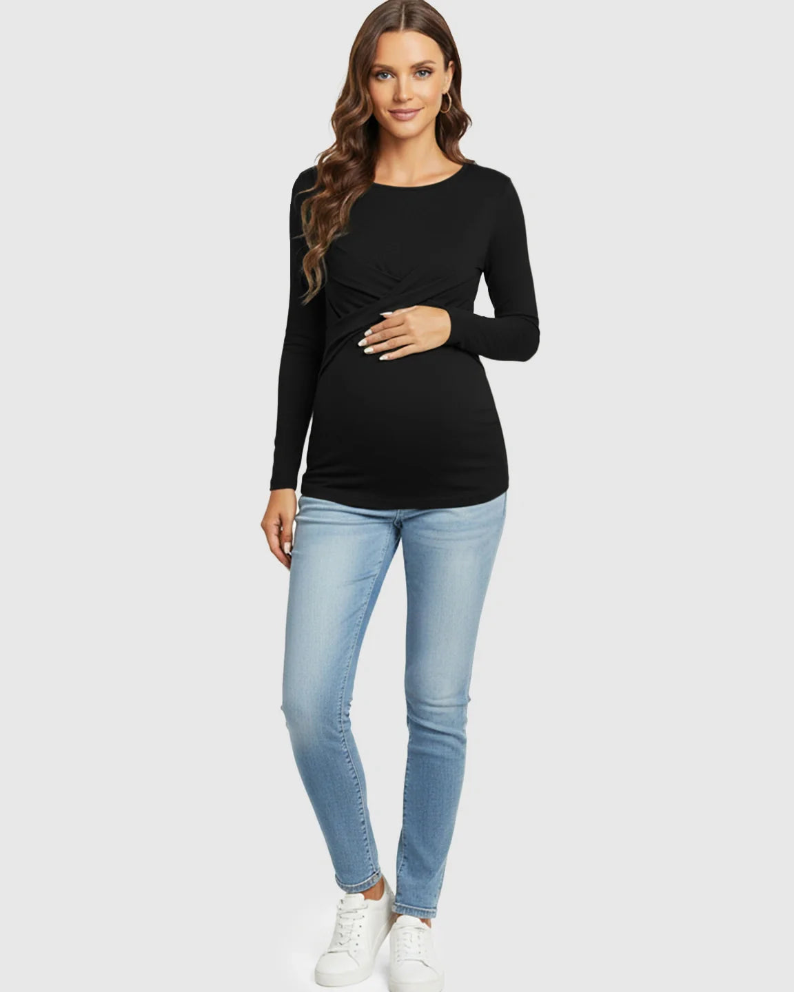 Maternity model in black cross-pleat fitted long sleeve top, light wash jeans, and white sneakers.