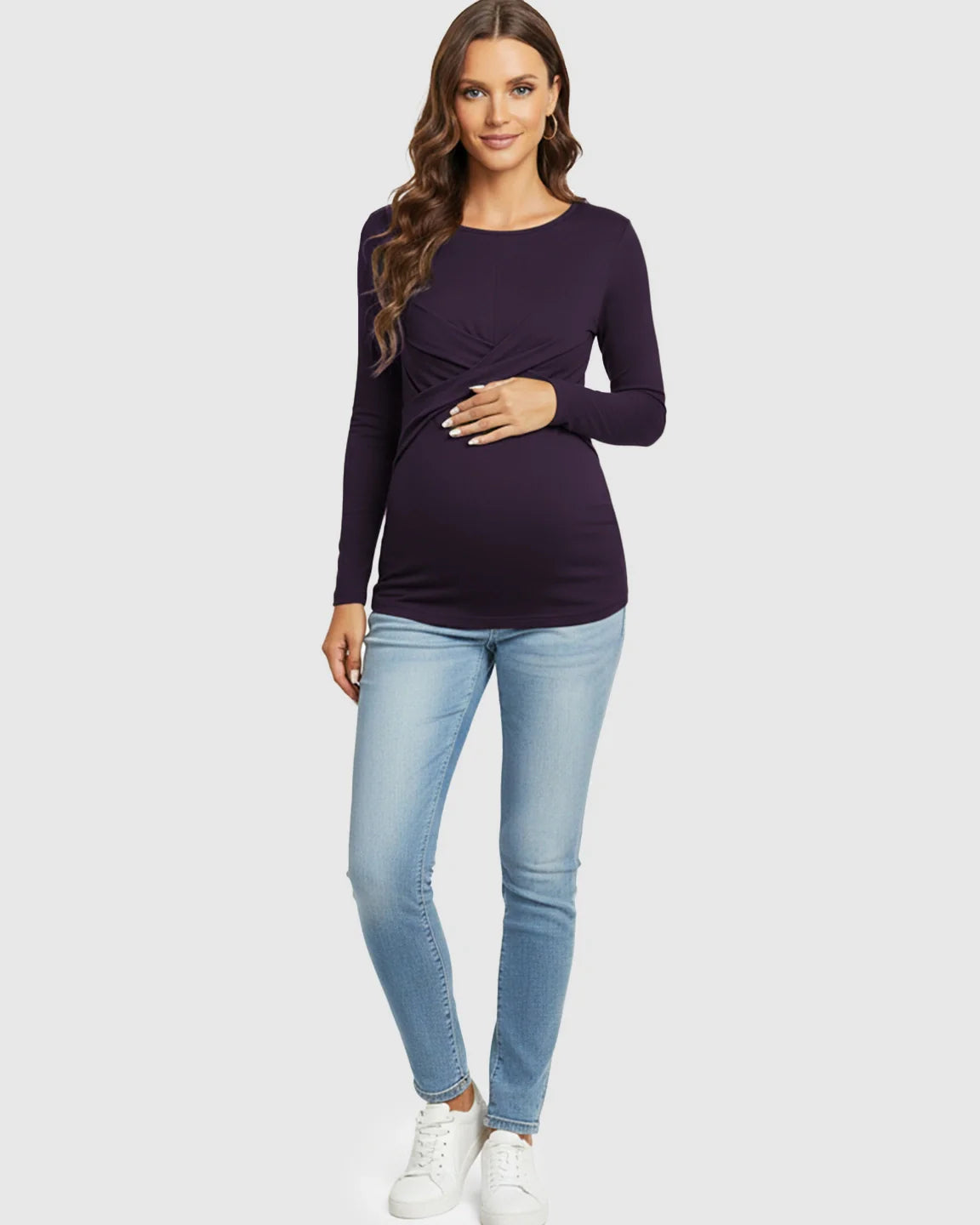 Pregnant woman in a purple cross-pleat fitted long sleeve maternity top, light wash jeans, and white sneakers.