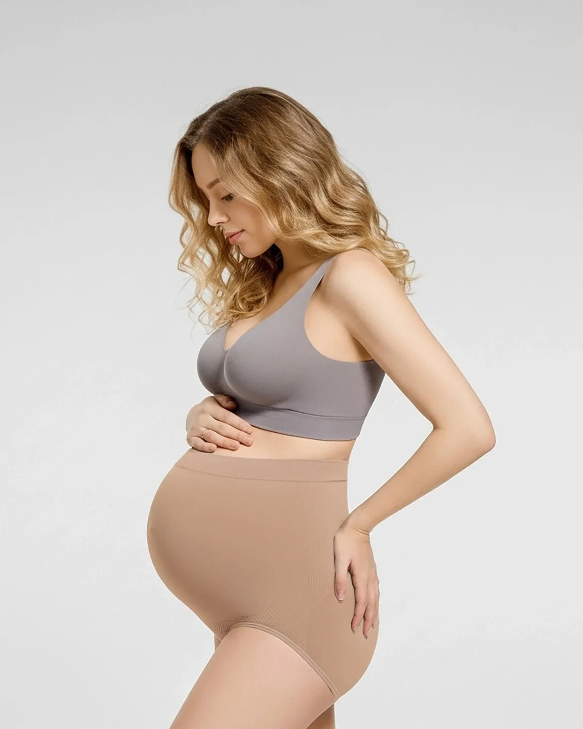Pregnant woman wearing seamless nude high-waist maternity panties and a gray bra, supporting her bump.