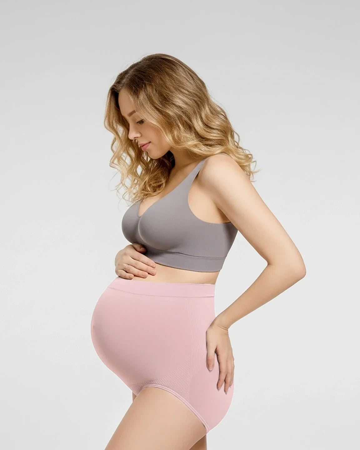 Pregnant woman in a gray bra and pink seamless high-waist maternity panties, gently cradling her bump.