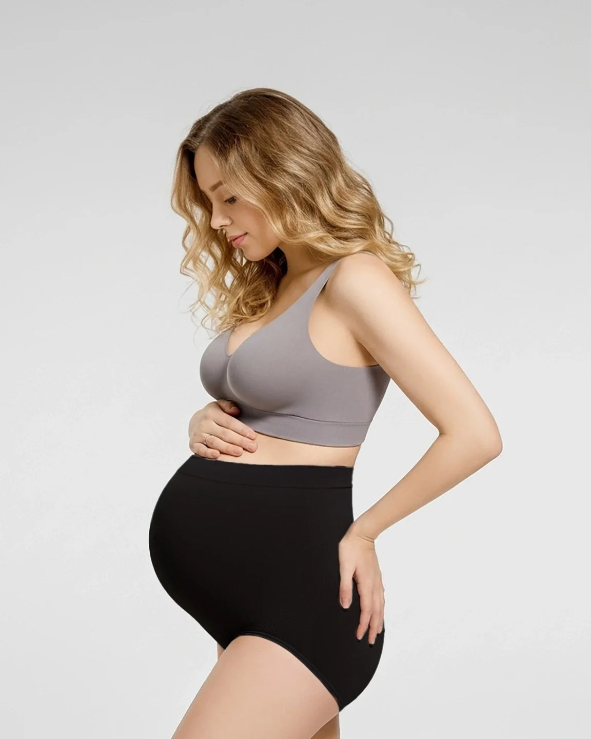 Pregnant woman in black high-waist seamless maternity panties and a gray bra, cradling her bump.