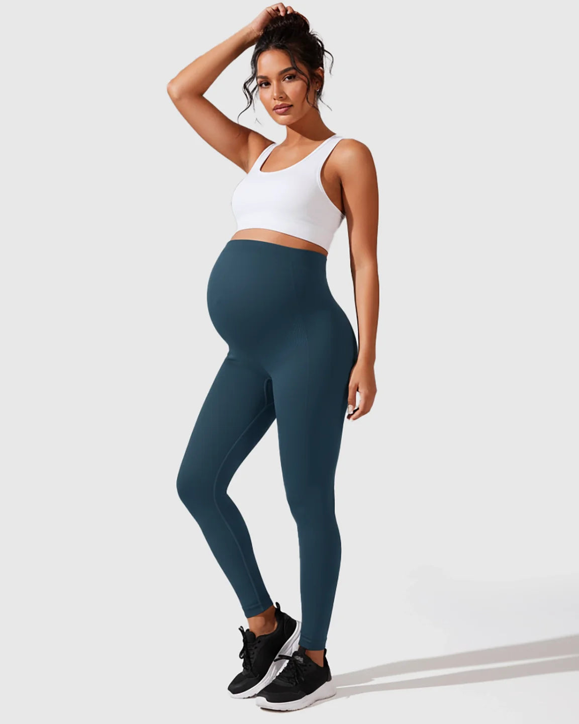 Pregnant woman in teal seamless high-waisted maternity leggings and a white sports bra, posing confidently.