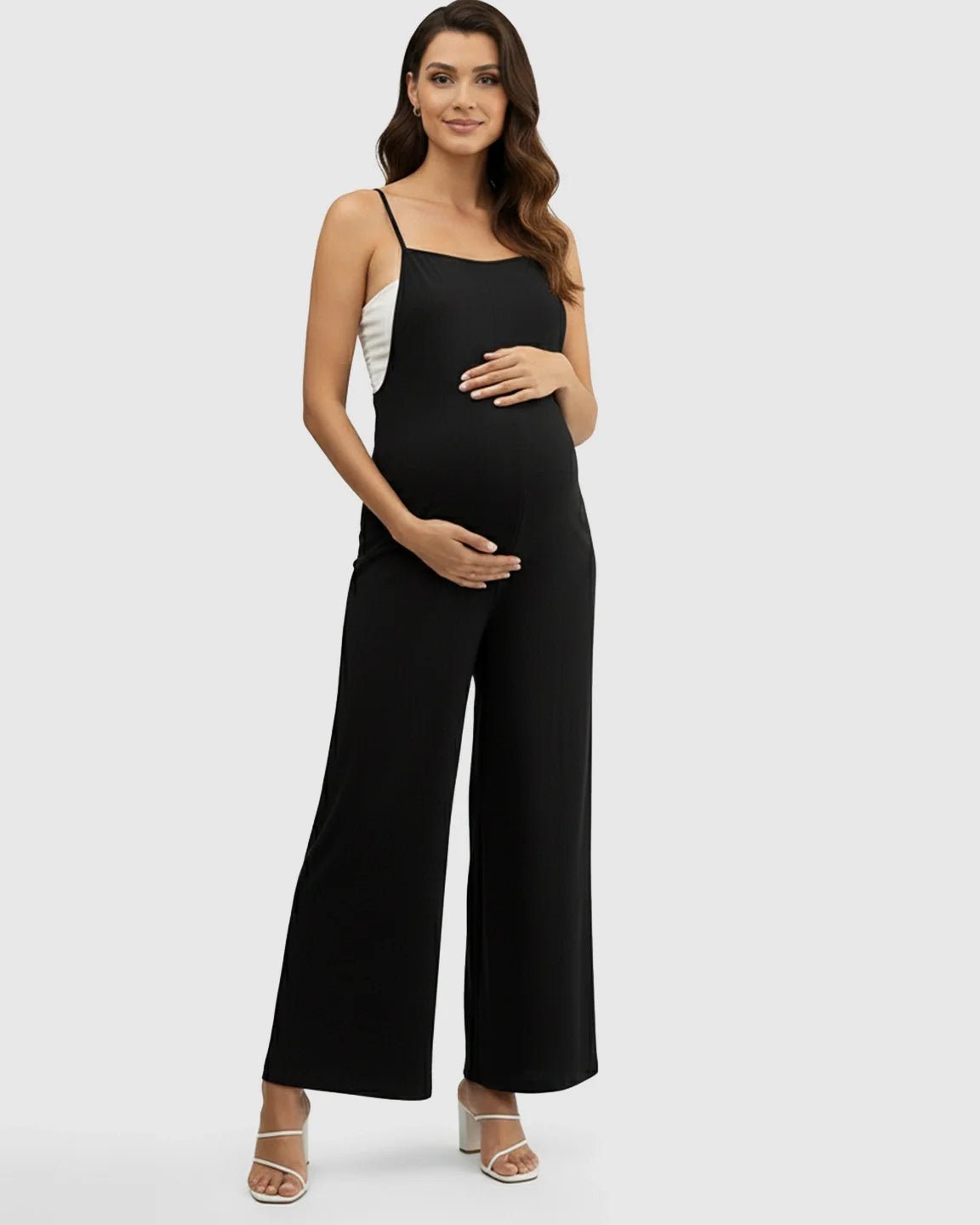 Pregnant model in a black spaghetti strap wide-leg maternity jumpsuit, hands on belly.