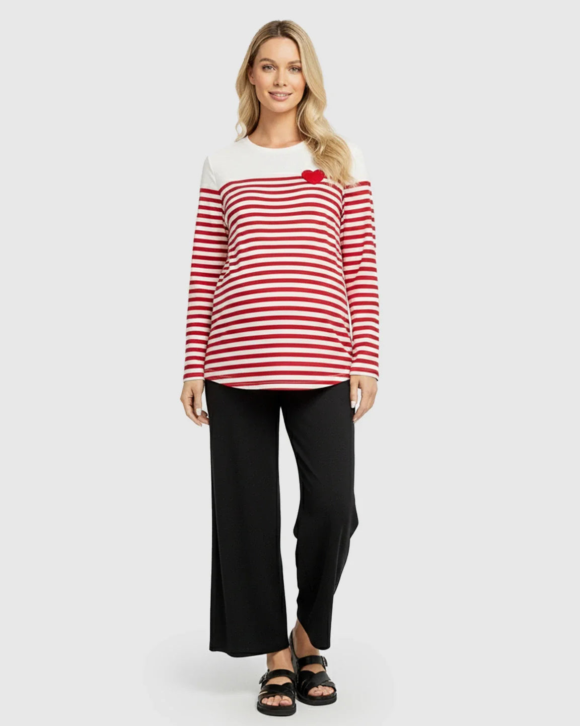 Blonde woman in a red and white striped maternity top with a red heart, black pants, and sandals.