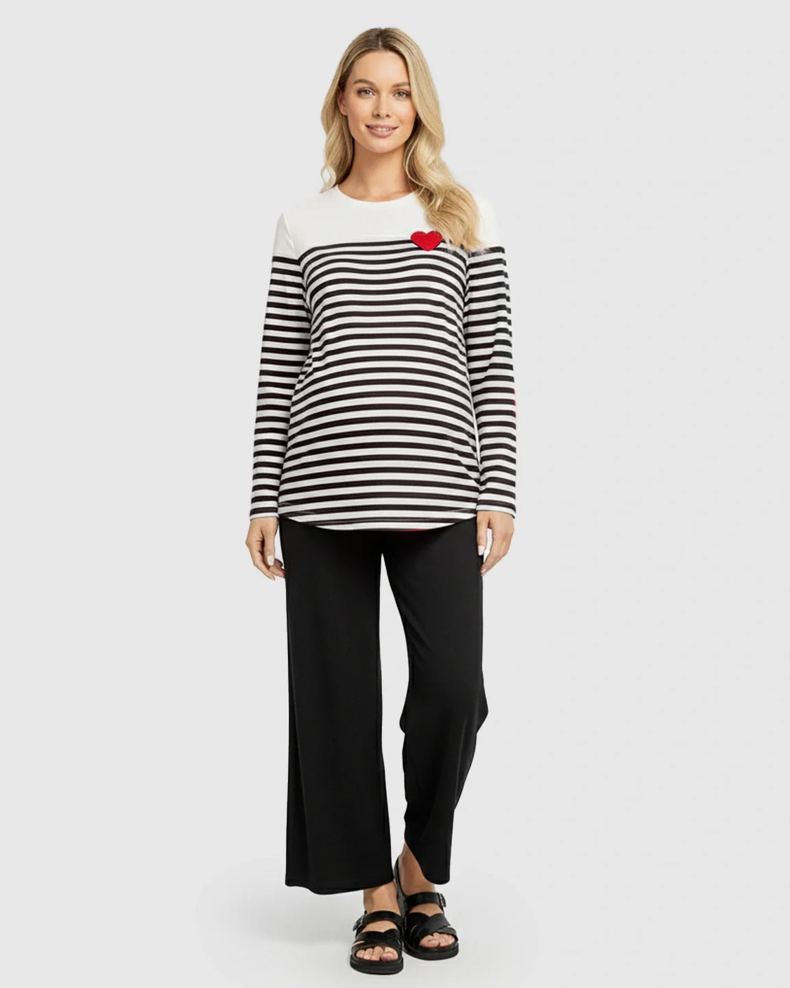 Blonde woman wearing a black and white striped long-sleeve maternity top with a red heart, and black pants.